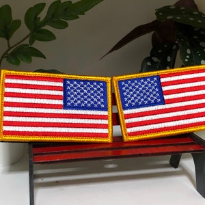 Puede incluir: Dos parches rectangulares bordados con la bandera estadounidense. Cada parche tiene un cantón azul con estrellas blancas, rayas rojas y blancas y un borde dorado. Los parches se muestran uno al lado del otro.