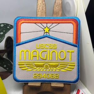 Puede incluir: Parche bordado de forma rectangular, con una estrella y el texto "USCSS MAGINOT" en amarillo. El parche tiene un borde azul y un diseño con acentos naranjas y rojos. El número "294062" está en la parte inferior.