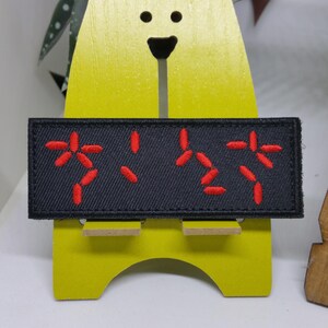 Puede incluir: Un parche rectangular negro con caracteres japoneses rojos bordados. El parche est&aacute; adherido a una figura de madera amarilla con forma de oso. Los caracteres japoneses est&aacute;n estilizados y parecen ser una forma de escritura o un s&iacute;mbolo.