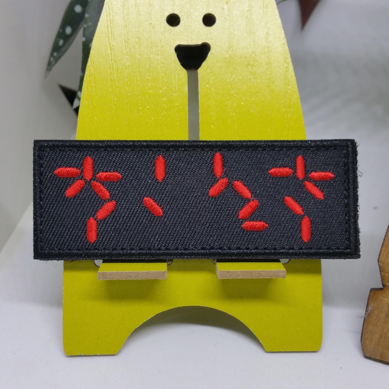 Puede incluir: Un parche rectangular negro con caracteres japoneses rojos bordados. El parche est&aacute; adherido a una figura de madera amarilla con forma de oso. Los caracteres japoneses est&aacute;n estilizados y parecen ser una forma de escritura o un s&iacute;mbolo.