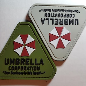 Peut inclure: Deux patchs triangulaires avec le logo d'Umbrella Corporation. Le logo est un parapluie rouge et blanc avec le texte "Umbrella Corporation" et la citation "Notre activité est la vie elle-même." Les patchs sont vert et gris.