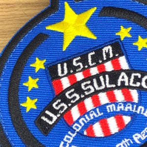 Puede incluir: Parche bordado con fondo azul, estrellas amarillas y el texto "U.S.C.M. U.S.S. SULACO COLONIAL MARINE". El parche presenta un diseño de escudo con rayas rojas y blancas.