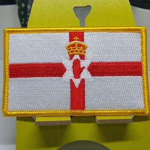 Puede incluir: Un parche blanco bordado con una cruz roja y una mano roja en el centro. La cruz está bordeada de oro y el parche tiene un borde dorado.