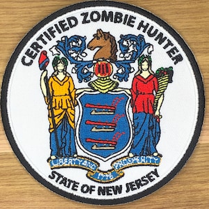 May include: A round patch with the words "CERTIFIED ZOMBIE HUNTER" encircling the New Jersey state seal. The seal includes figures, a horse, and the motto "LIBERTY AND PROSPERITY" and "STATE OF NEW JERSEY".