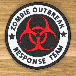 Könnte beinhalten: Runder Aufnäher mit dem Text "ZOMBIE OUTBREAK RESPONSE TEAM" in Weiß. Er hat einen schwarzen Hintergrund mit einem roten Gefahrensymbol in der Mitte und zwei schwarzen Sternen.