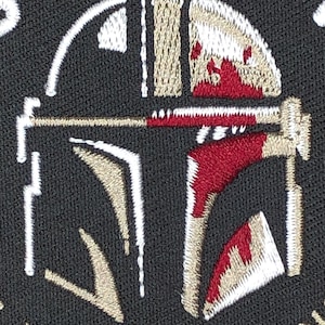 Könnte beinhalten: Gestickter Aufnäher mit einer detaillierten Darstellung eines Mandalorianer-Helms. Das Design verwendet weiße, beige und rote Akzente auf schwarzem Hintergrund. Die ikonische Form des Helms ist deutlich definiert.