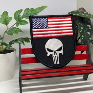 May include: A black and white embroidered patch with an American flag and a skull. The patch is on a red wooden bench.