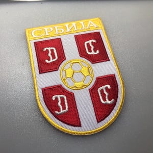 May include: Embroidered patch featuring the Serbian national football team crest. The crest is a white shield with a red cross and four red cyrillic letters "C" in each quadrant. A gold soccer ball is in the center of the cross. The shield is outlined in gold and the text "SRBIJA" is above the shield.