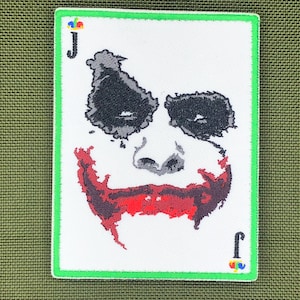 Könnte beinhalten: Gestickter Aufnäher mit dem Gesicht des Jokers aus dem Batman-Franchise. Das Design umfasst schwarzes Augen-Make-up, ein rotes Lächeln und den Buchstaben "J" in den Ecken. Der Aufnäher hat einen weißen Hintergrund mit einem grünen Rand.