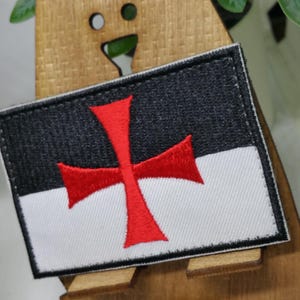 Puede incluir: Un parche bordado blanco y negro con una cruz roja. La cruz es un s&iacute;mbolo de los Caballeros Templarios.