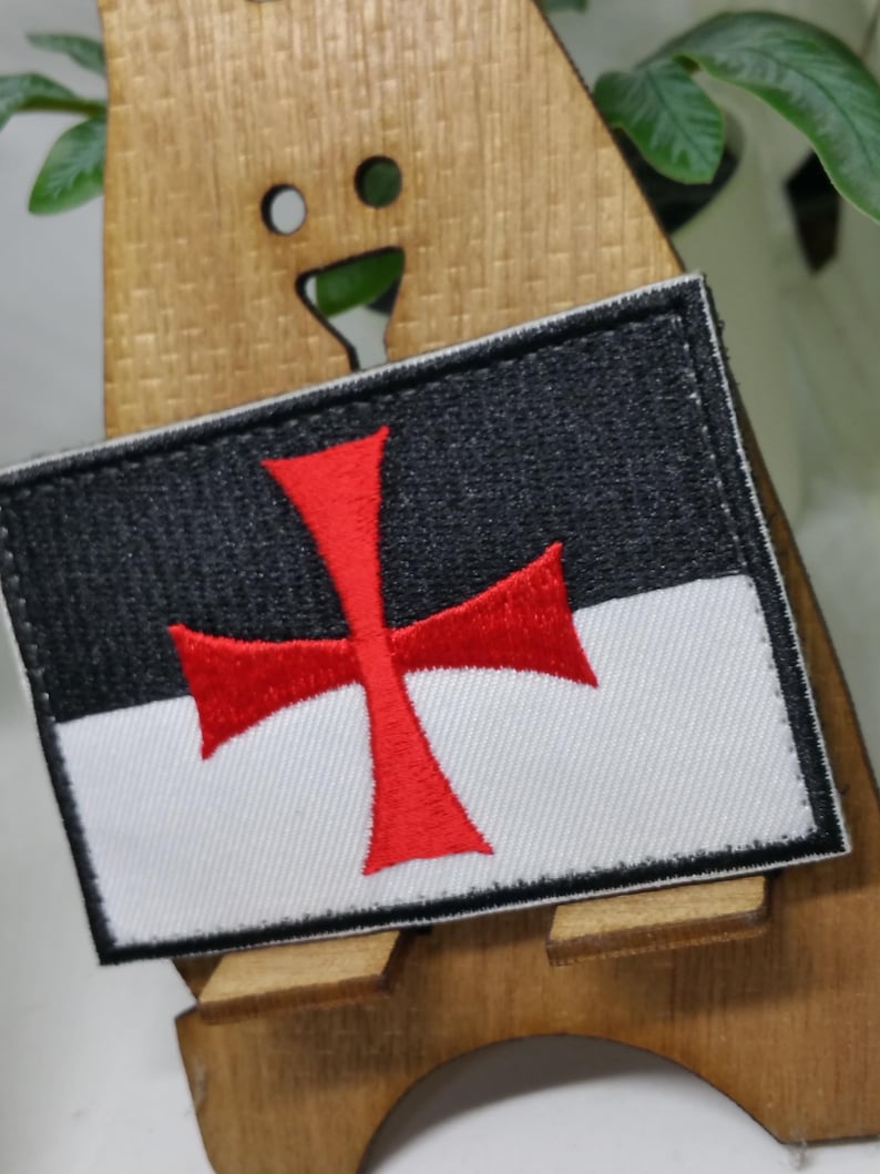 Puede incluir: Un parche bordado blanco y negro con una cruz roja. La cruz es un s&iacute;mbolo de los Caballeros Templarios.