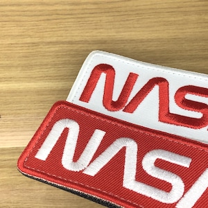Könnte beinhalten: Zwei gestickte NASA-Patches. Ein Patch hat einen weißen Hintergrund mit roten Nähten, und der andere hat einen roten Hintergrund mit weißen Nähten. Das NASA-Logo ist auf beiden Patches deutlich sichtbar.