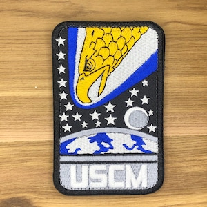 May include: Embroidered patch featuring a yellow eagle head with a blue and white background, set against a black starry sky. The patch includes the letters "USCM" in white. The patch is rectangular with rounded corners.