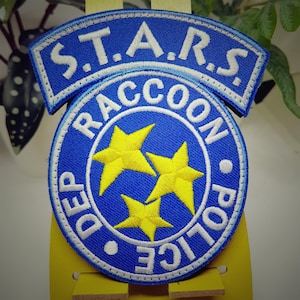 May include: A blue and white embroidered patch with the text "S.T.A.R.S. Raccoon Police Dep." and three yellow stars.