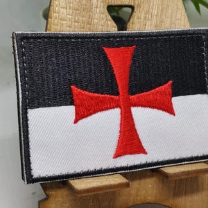 Puede incluir: Un parche bordado en blanco y negro con una cruz roja. El parche es una representaci&oacute;n de los Caballeros Templarios.