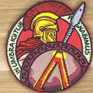May include: Embroidered patch depicting a Spartan warrior in a yellow helmet and red armour, holding a spear and shield. The patch has a red and tan background with the text "IN UMBRA IGITUR ARENABIMUS" around the edge.