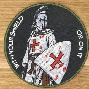 Op de afbeelding: Geborduurde patch met een ridder in harnas met een rood kruis op zijn borst en schild. De patch heeft de tekst "WITH YOUR SHIELD OR ON IT" langs de rand. De ridder staat tegen een zwarte achtergrond met een groene rand.
