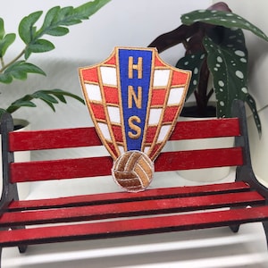 Peut inclure: Un banc en bois rouge avec un écusson brodé bleu, blanc et or avec les lettres "HNS" et un ballon de football.