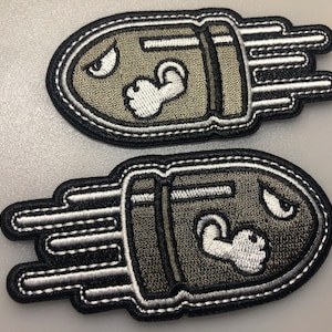 May include: Two embroidered bullet-shaped patches. They have a grey body, white detailing, and a black border. Each patch features a cartoon face with an angry expression and a flexing arm.