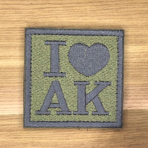 Può includere: Toppa quadrata con la scritta "I ♥ AK" in lettere grigie su sfondo verde. La toppa ha un bordo grigio ed è probabilmente un articolo ricamato.