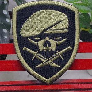 Puede incluir: Un parche bordado verde y negro con una calavera que lleva una boina y dagas cruzadas. El parche tiene forma de escudo.