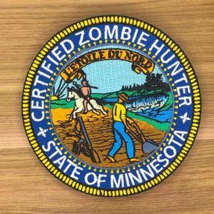 Könnte beinhalten: Runder, gestickter Aufnäher mit der Aufschrift "CERTIFIED ZOMBIE HUNTER" und "STATE OF MINNESOTA". Die Mitte zeigt eine Landschaft mit Figuren und dem Satz "L'ETOILE DU NORD". Der Rand ist gelb.