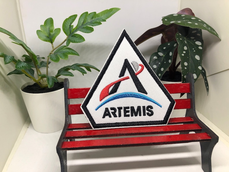 May include: A white triangular patch with a black stylised letter A, a red and blue crescent moon, and the word "ARTEMIS" in black text.