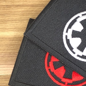 Może przedstawiać: Dwie czarne, haftowane naszywki z motywem Star Wars. Jedna naszywka przedstawia biały symbol imperialny, a druga czerwony symbol imperialny. Naszywki są kwadratowe i mają czarną obwódkę.
