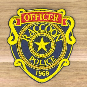May include: Embroidered patch featuring a police badge design. The patch is navy blue with yellow and red accents. The text "OFFICER" is in red, and "RACCOON POLICE 1969" is in yellow.
