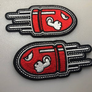 May include: Two red and white embroidered iron-on patches shaped like a bullet with a cartoon face and a fist. The patches have a black outline and white stitching.