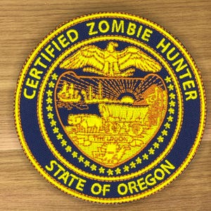 Puede incluir: Parche circular con el texto "CERTIFIED ZOMBIE HUNTER STATE OF OREGON" en letras amarillas sobre fondo azul marino. El parche presenta un águila dorada, una carreta y un diseño de puesta de sol.