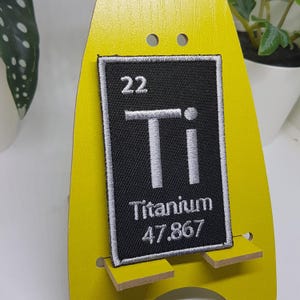 Może przedstawiać: Czarno-biała haftowana łatka z elementem tytanu z układu okresowego. Łatka przedstawia symbol elementu "Ti", liczbę atomową "22" i masę atomową "47,867".