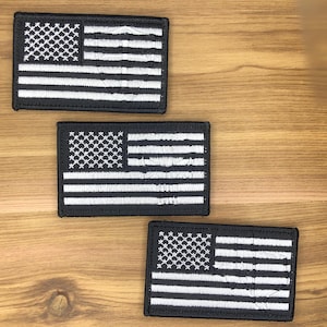 May include: Three black and white embroidered American flag patches. Each patch features a field of white stars in the upper left corner and white stripes on a black background. The patches are rectangular and have a black border.