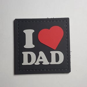 Può includere: Toppa nera con cuciture bianche attorno ai bordi. La toppa ha una "I" e un "DAD" bianchi con un cuore rosso in mezzo. Il cuore è pieno di rosso.