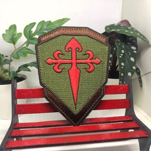 Puede incluir: Un parche bordado en forma de escudo con una cruz roja y una espada sobre un fondo verde y marrón.