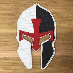 May include: Embroidered patch in the shape of a Spartan helmet, split vertically into white and black sections. A red cross design is centered on the helmet. The patch is likely for clothing or accessories.