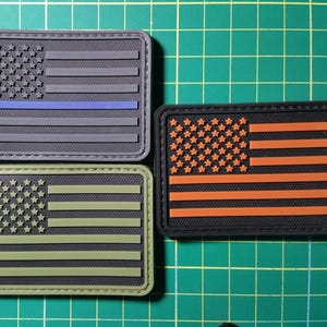 May include: Three American flag patches, each with a different colored stripe. The first patch has a blue stripe, the second has an orange stripe, and the third has a green stripe.