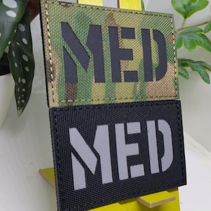 Pode incluir: Duas etiquetas retangulares, uma camuflada e outra preta, ambas exibindo a palavra "MED" em letras de bloco em negrito. A etiqueta camuflada tem um padrão verde, marrom e bege. A etiqueta preta tem letras cinzas.