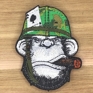 May include: Embroidered patch featuring a monkey wearing a camouflage helmet and smoking a cigar. The monkey's face is white with a black beard and furrowed brow. An ace of spades card is visible on the helmet.