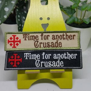 Pu&ograve; includere: Due toppe ricamate con il testo "Time for another Crusade" e un simbolo di croce rossa. La toppa superiore &egrave; beige con testo marrone e la toppa inferiore &egrave; nera con testo bianco.
