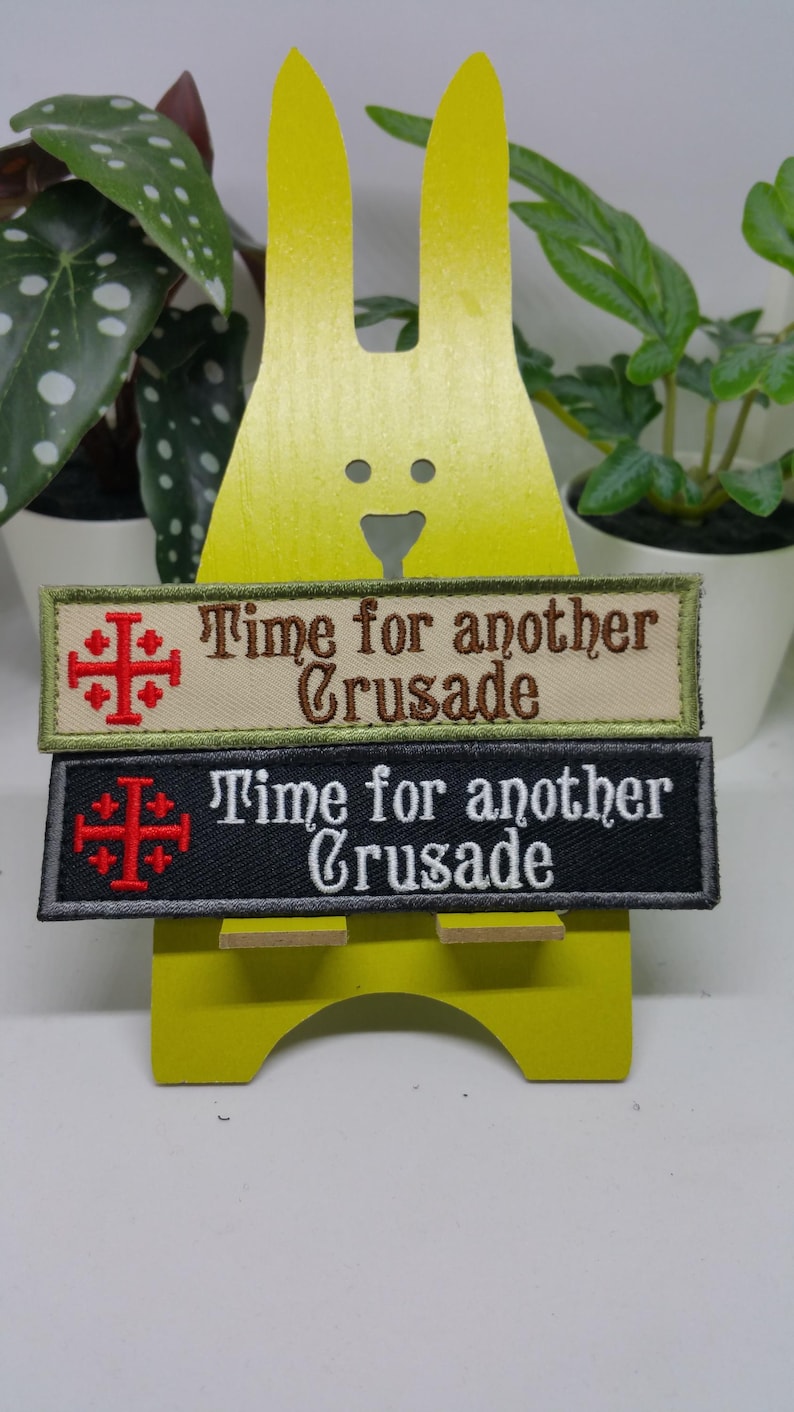 Pu&ograve; includere: Due toppe ricamate con il testo "Time for another Crusade" e un simbolo di croce rossa. La toppa superiore &egrave; beige con testo marrone e la toppa inferiore &egrave; nera con testo bianco.