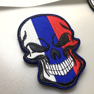 May include: A white, blue and red embroidered skull patch with a black outline. The skull is smiling and has a black eye.