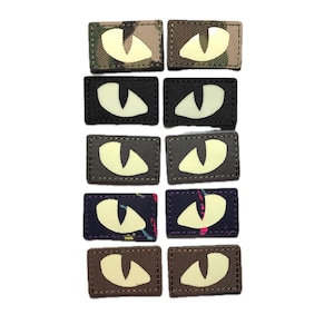 Op de afbeelding: Een verzameling rechthoekige patches met gloeiende oogontwerpen. De patches hebben verschillende achtergrondpatronen, waaronder camouflage, zwart en stoffen met patronen. De ogen gloeien met een heldere, gebroken witte kleur.