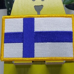 May include: A white and blue embroidered patch with a yellow border, depicting the flag of Finland.