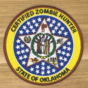 May include: A circular patch with the text "CERTIFIED ZOMBIE HUNTER" and "STATE OF OKLAHOMA". The patch features a blue star with white stars, and a central design with figures and a wreath. The border is yellow and brown.
