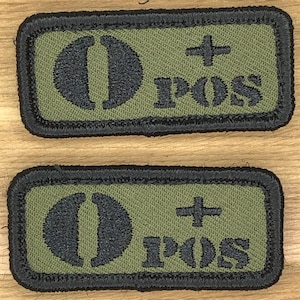 Può includere: Due toppe rettangolari con la scritta "O+ POS" ricamata in nero su sfondo verde oliva. Le toppe hanno un bordo nero.