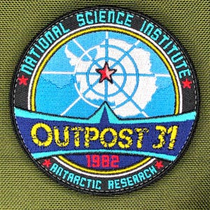 Może przedstawiać: Okrągła, haftowana naszywka z napisem "NATIONAL SCIENCE INSTITUTE" i "OUTPOST 31 ANTARCTIC RESEARCH 1982". Naszywka przedstawia mapę Antarktydy w kolorze niebiesko-białym z czerwoną gwiazdą pośrodku.