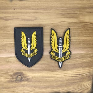 Könnte beinhalten: Zwei gestickte Aufnäher mit einem gelben, geflügelten Dolch-Design auf schwarzem Hintergrund. Der Text "WHO DARES WINS" ist auf einem gelben Banner zu sehen. Ein Aufnäher ist schildförmig, der andere hat Flügel.