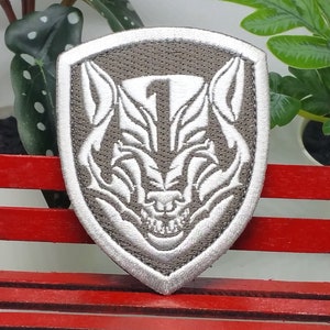 Puede incluir: Un parche bordado gris y blanco con una cabeza de lobo y el número "1" dentro de una forma de escudo.