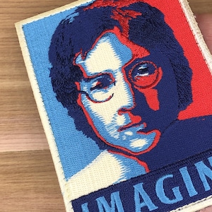 Può includere: Toppa ricamata raffigurante un ritratto di uomo con occhiali rotondi, nei toni del blu e del rosso. La parola "IMAGIN" è parzialmente visibile in basso. La toppa ha un bordo bianco ed è destinata ai tessuti.